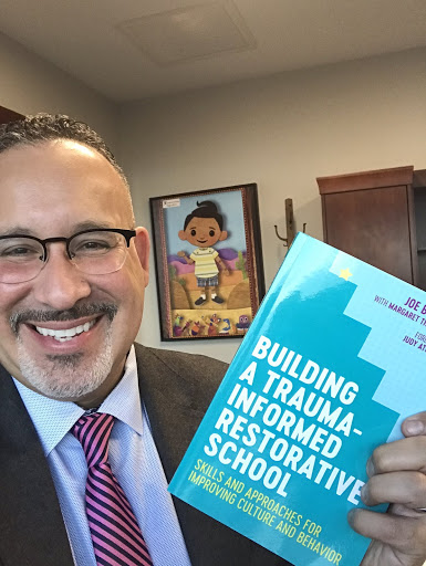 Miguel Cardona holds a book with the the title "Building a Trauma-Informed Restorative School