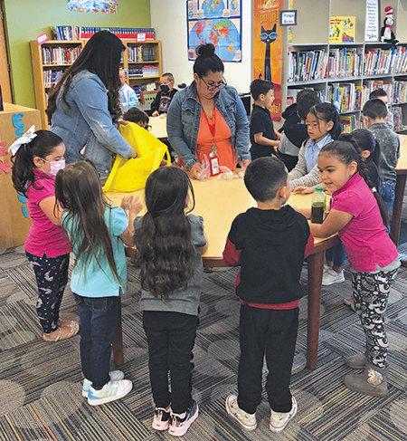 elementary students stand around a table in a classroom which a teacher instructs them