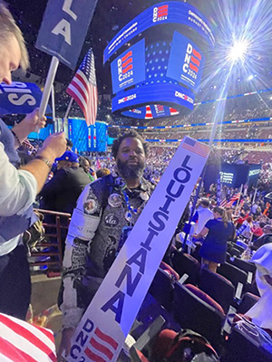 A man stands at a convention, holding a sign that reads Louisiana, and is surrounded by attendees.