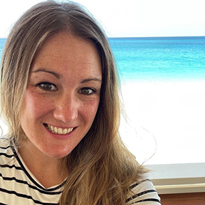 A woman wearing a striped shirt smiles warmly at the camera, with beach and ocean water set behind her.