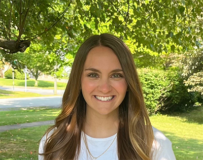 A woman in a white t-shirt poses with a smile against a backdrop of trees.