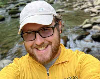 A man with glasses, smiling, and wearing yellow shirt takes a selfie.