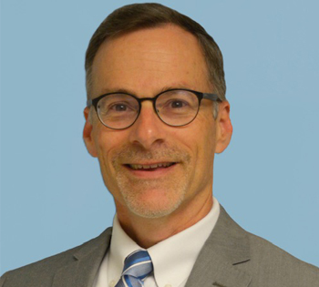 A man smiling and wearing glasses dressed in a suit and tie looks directly to camera.