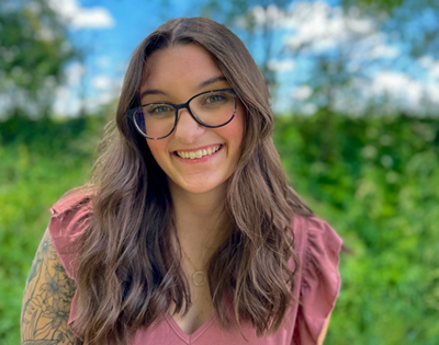 A tattooed woman wearing glasses stands in front of a green field of trees.