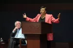 Becky Pringle speaks at a town hall while Senator Bernie Sanders sits on stage and listens