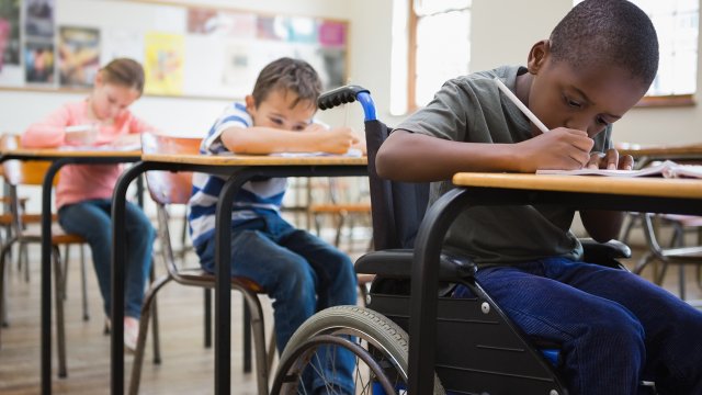 African American boy in wheelchair writing at school desk with classmates in background