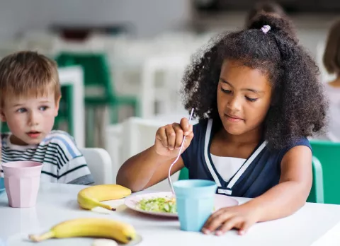 Young girl of color eating lunch while a young boy looks at her.