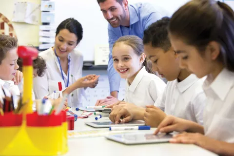 A group of school children can be seen working on digital tablets, two teachers can be seen behind them helping and supervising