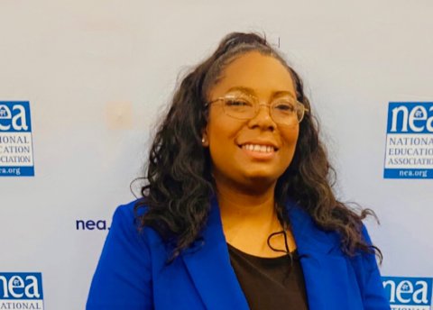 A woman in a blue blazer stands confidently in front of an NEA banner.