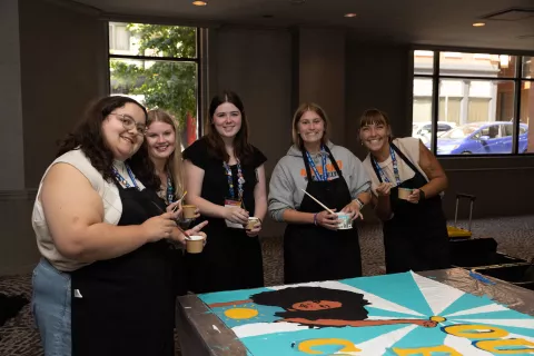 Aspiring Educators stand smiling with paintbrushes in their hands, working on an art activism piece.