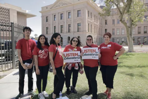 Educators in red shirts stand outside the Arizona capitol holding signs.