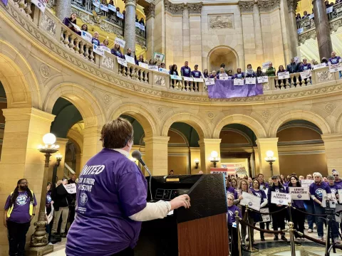 An educator in a purple shirt speaks to a crowded state capital.